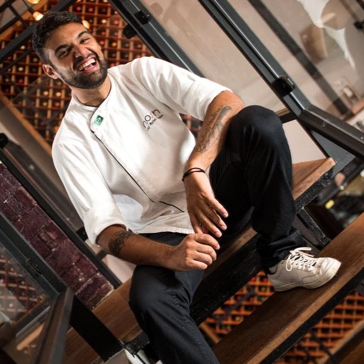 Chef Rafael Madeira sorrindo na escada do NOAH, com a adega ao fundo